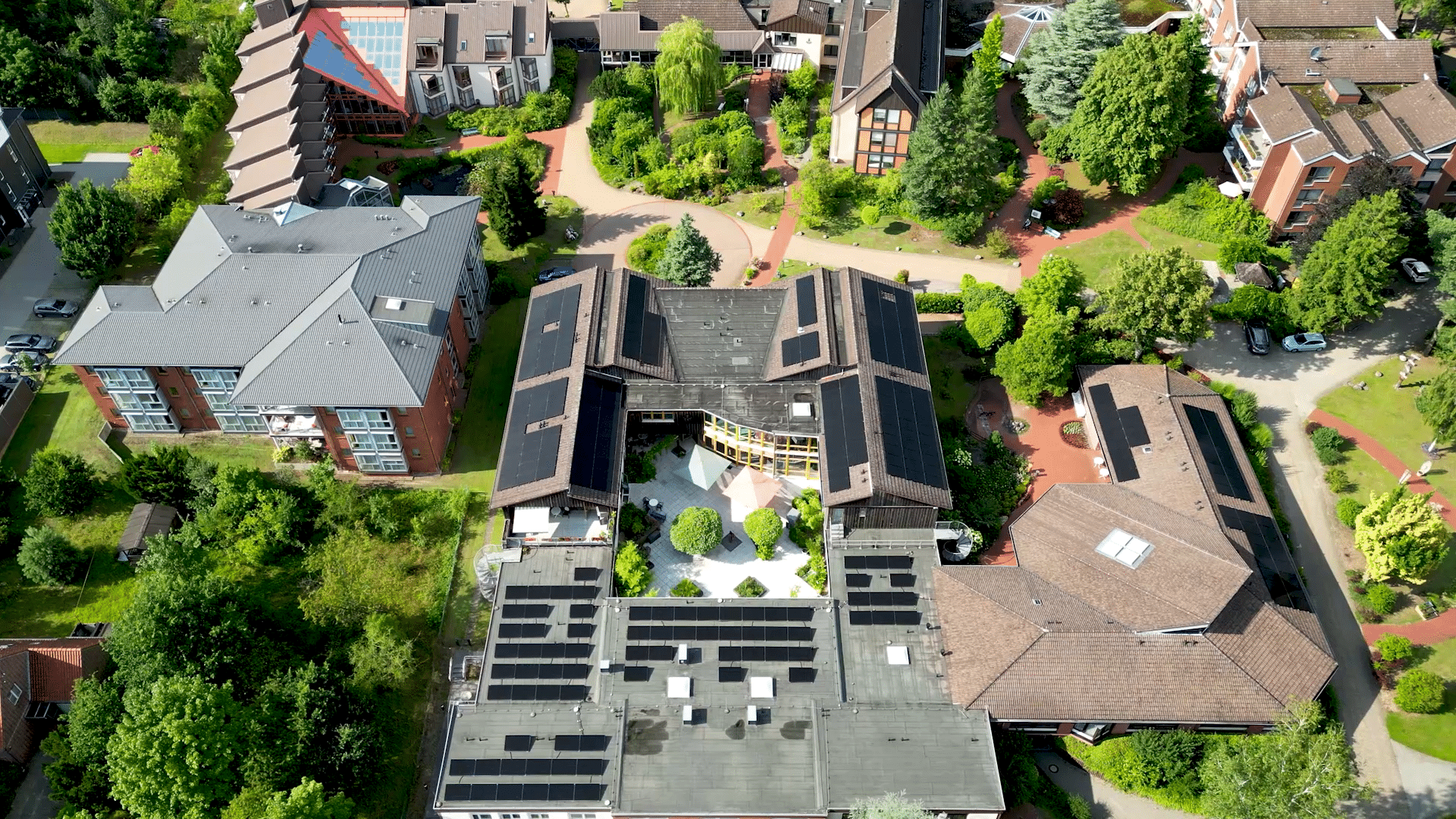 Senkrechte Luftaufnahme des Innenhofgebäudes in Soltau, Photovoltaikmodule in Reihen auf der Flachdachfläche und in Feldern auf den angrenzenden Ziegeldächern, im Innenhof eine Terrasse mit Sonnenschirmen