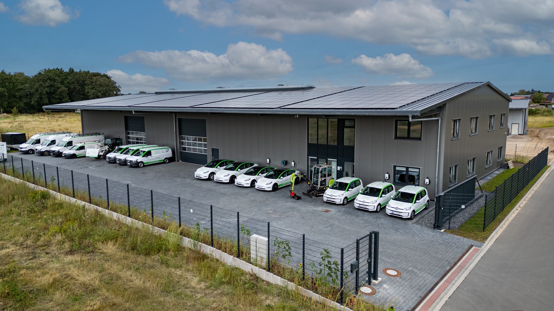 Schräge Luftaufnahme der langen Halle. Auf der Dachfläche liegen dunkle Photovoltaikmodule in dichten Reihen, im Hof davor stehen weiße Transporter und Pkw nebeneinander.