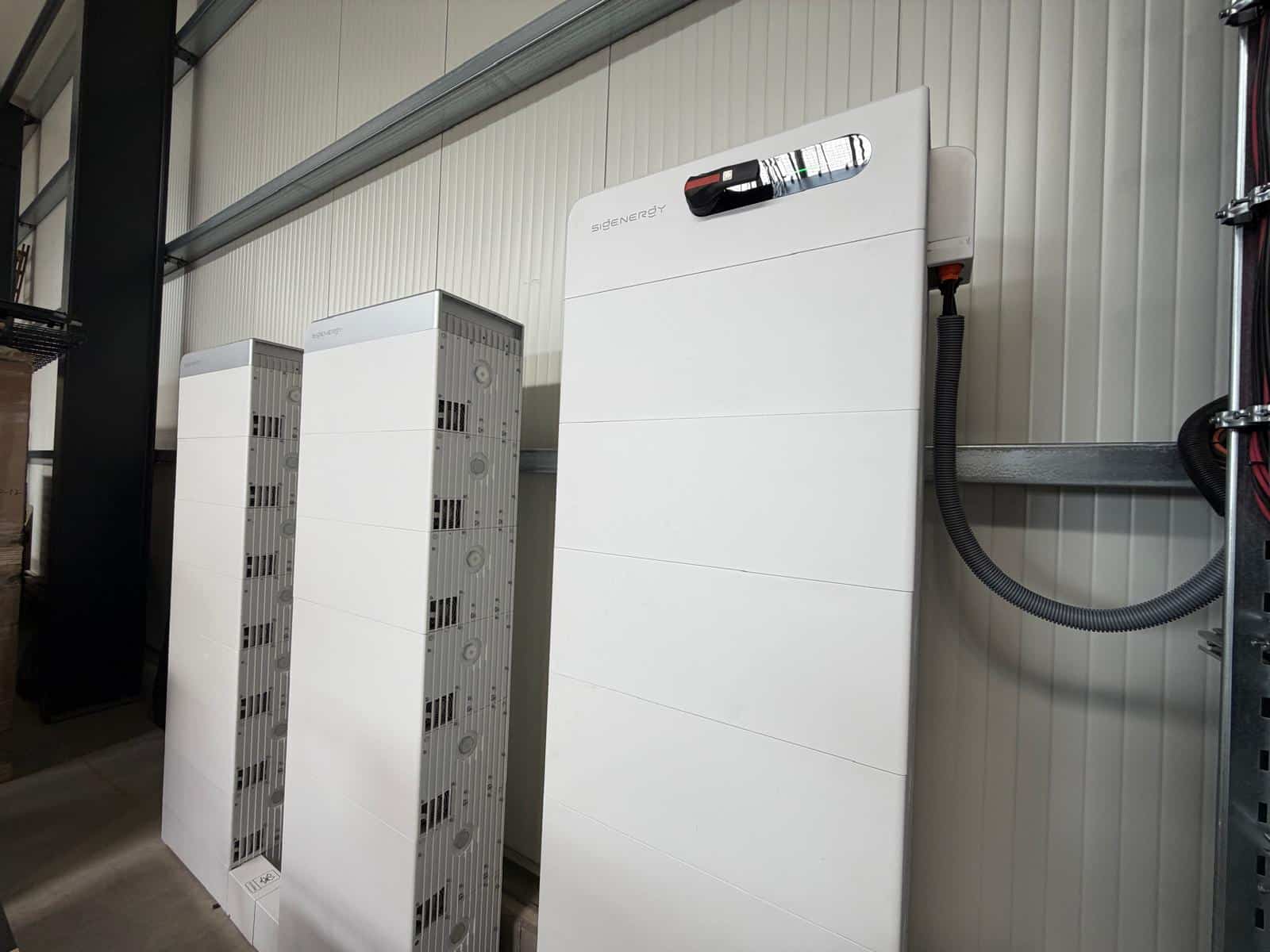 Drei weiße Batterietürme stehen nebeneinander an einer hellen Hallenwand. Der vordere trägt oben eine schmale Anzeigeleiste, rechts führt ein graues Wellrohr zur Wandinstallation.