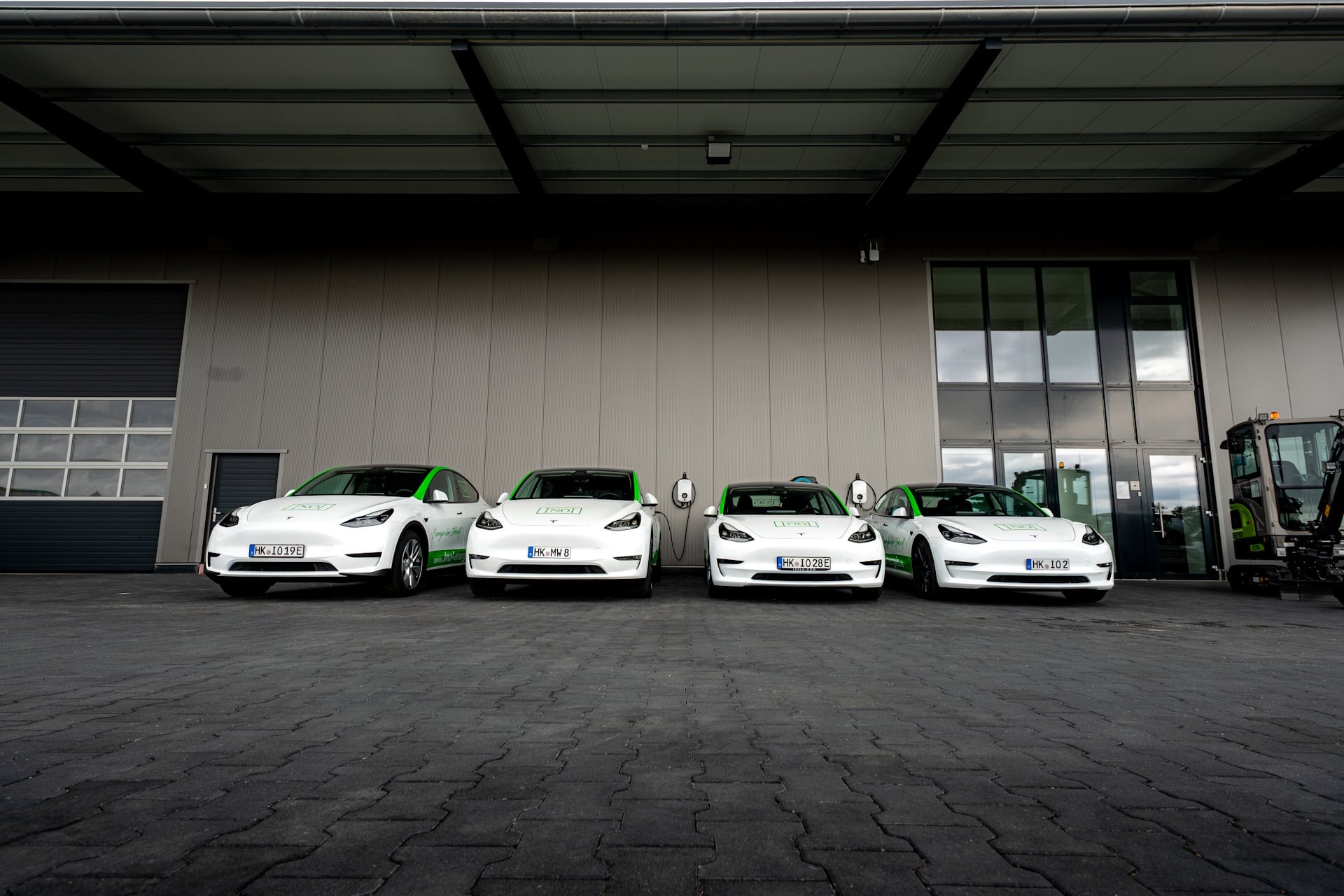 Vier weiße Teslas mit grüner Beschriftung stehen frontal nebeneinander unter einem Vordach. Zwischen den beiden mittleren Fahrzeugen hängt eine Wallbox an der Wand.