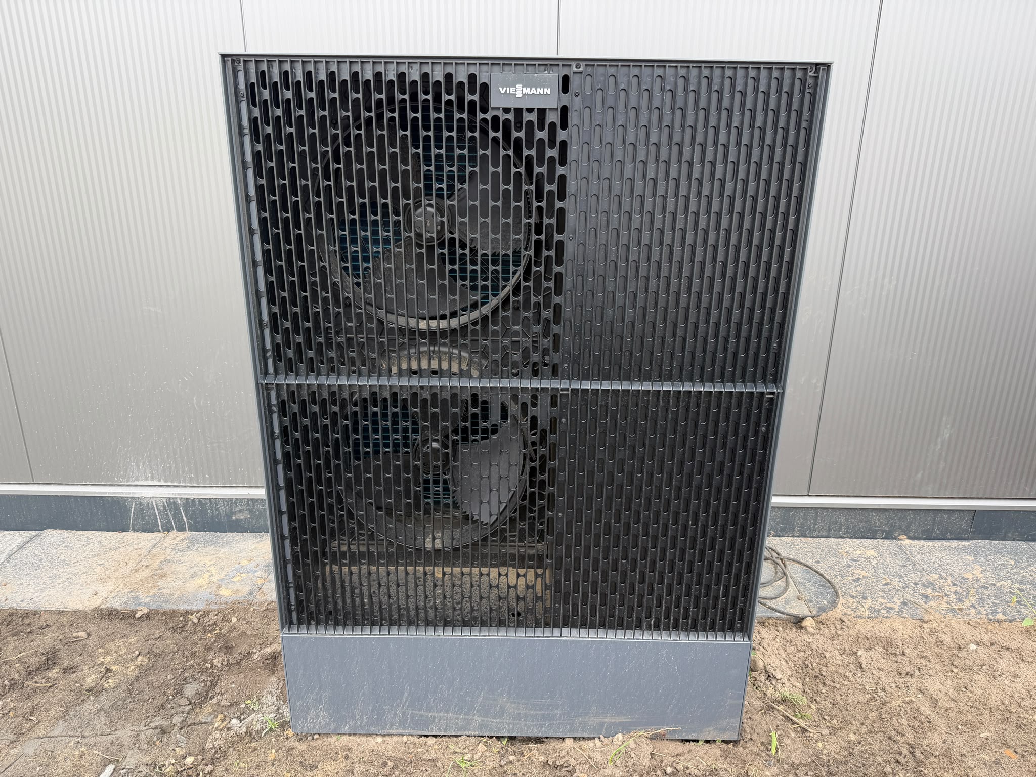 Das graue Außengerät einer Viessmann-Wärmepumpe steht auf Betonplatten vor einer hellen Fassade. Hinter dem dunklen Schutzgitter sind zwei große Ventilatoren übereinander zu erkennen.
