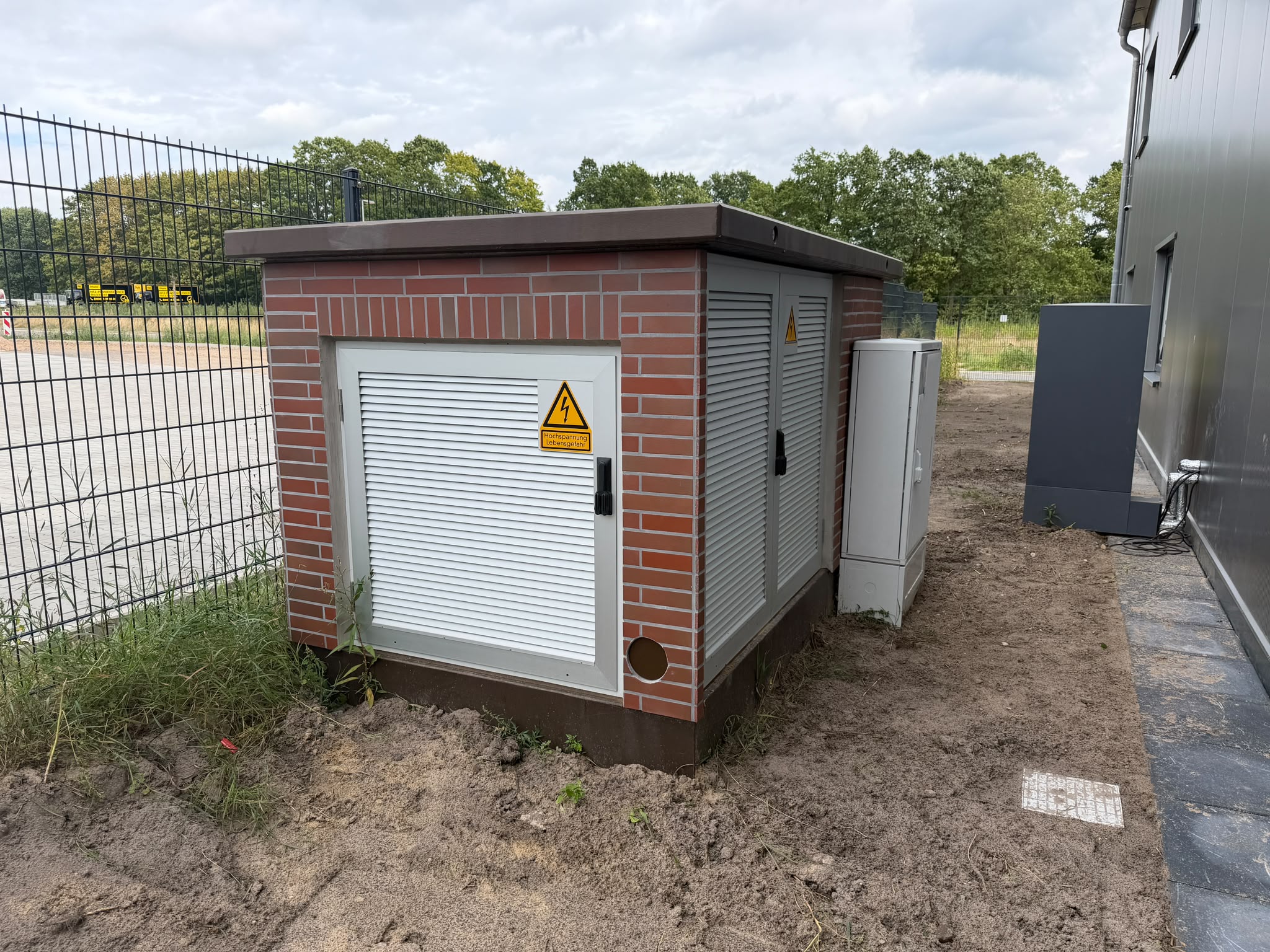 Eine gemauerte Trafostation mit rotem Klinker und hellen Lüftungstüren steht am Zaun. Ein gelbes Warnschild an der Tür weist auf Hochspannung und Lebensgefahr hin.