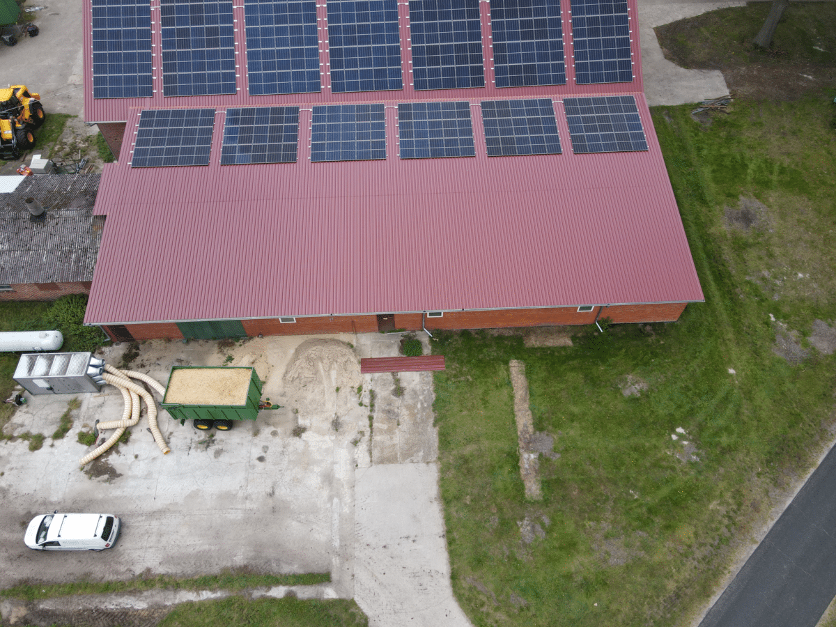 Draufsicht per Drohne auf eine Halle, deren obere Dachhälfte Photovoltaikmodule trägt, während die untere rote Dachfläche frei ist, davor Hof, Anhänger und Wiese.