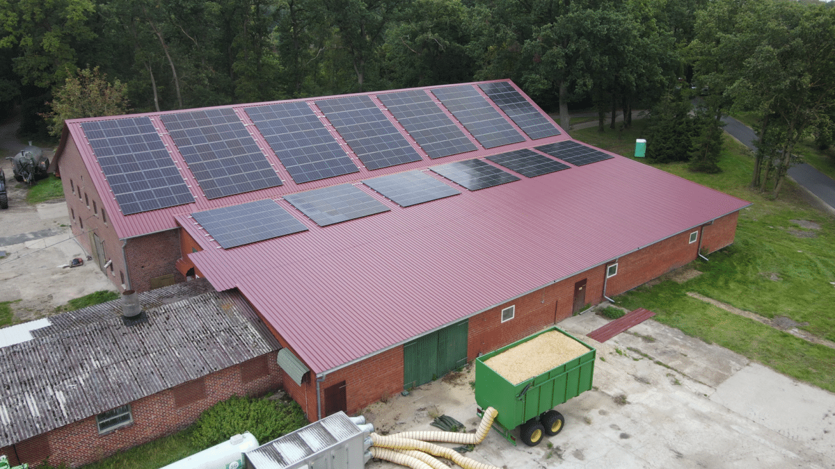 Schrägluftbild einer langen Backsteinhalle mit rotem Trapezblechdach und zwei Reihen Photovoltaikmodulen, davor auf dem Hof ein grüner Anhänger mit Holzhackschnitzeln und angeschlossenen gelben Luftschläuchen.