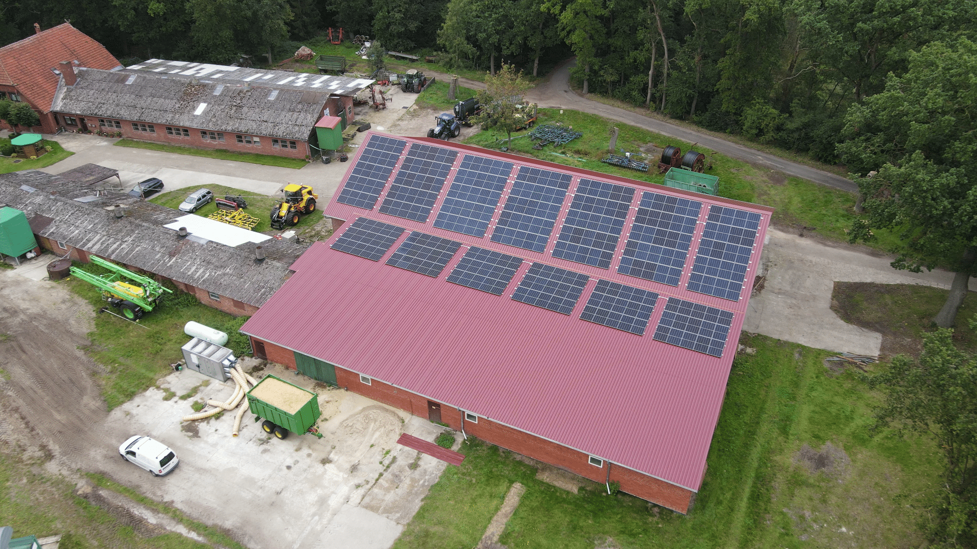 Schrägluftbild einer Halle mit rotem Trapezblechdach und zwei Reihen Photovoltaikmodulen, daneben ältere Nebengebäude, Landmaschinen, ein grüner Anhänger mit Luftschläuchen und Wald im Hintergrund.