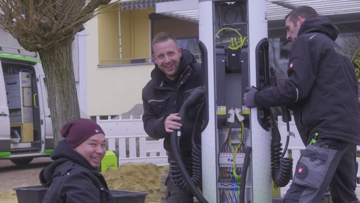 Drei Monteure der INOL GmbH bauen vor einem Gebäude eine Ladesäule für Elektroautos auf. Die Säule ist geöffnet, innen sind Schutzschalter, Klemmen und Kabel zu sehen. Zwei Mitarbeiter halten die Ladekabel und lachen in die Kamera, ein dritter kniet vorn neben einem Sandhaufen. Links steht ein weißer Transporter mit grünen Streifen und offener Seitentür. Solche Ladeinfrastruktur plant und installiert INOL mit eigenen Elektrikern.