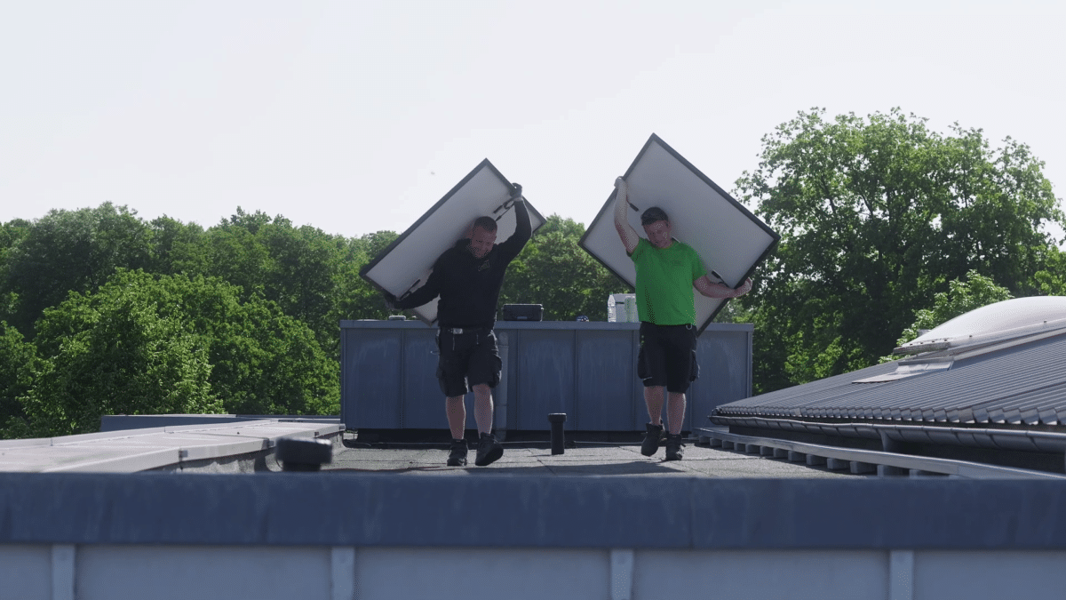 Zwei Monteure der INOL GmbH tragen Solarmodule für eine Photovoltaikanlage über ein Flachdach. Beide halten je ein großes Modul schräg über Kopf und Schulter und lächeln. Der linke trägt einen schwarzen Kapuzenpullover, der rechte ein grünes T-Shirt, beide kurze Arbeitshosen. Im Hintergrund stehen ein Dachaufbau und hohe Laubbäume unter hellem Himmel. Die Montage übernehmen bei INOL eigene, fest angestellte Handwerker.