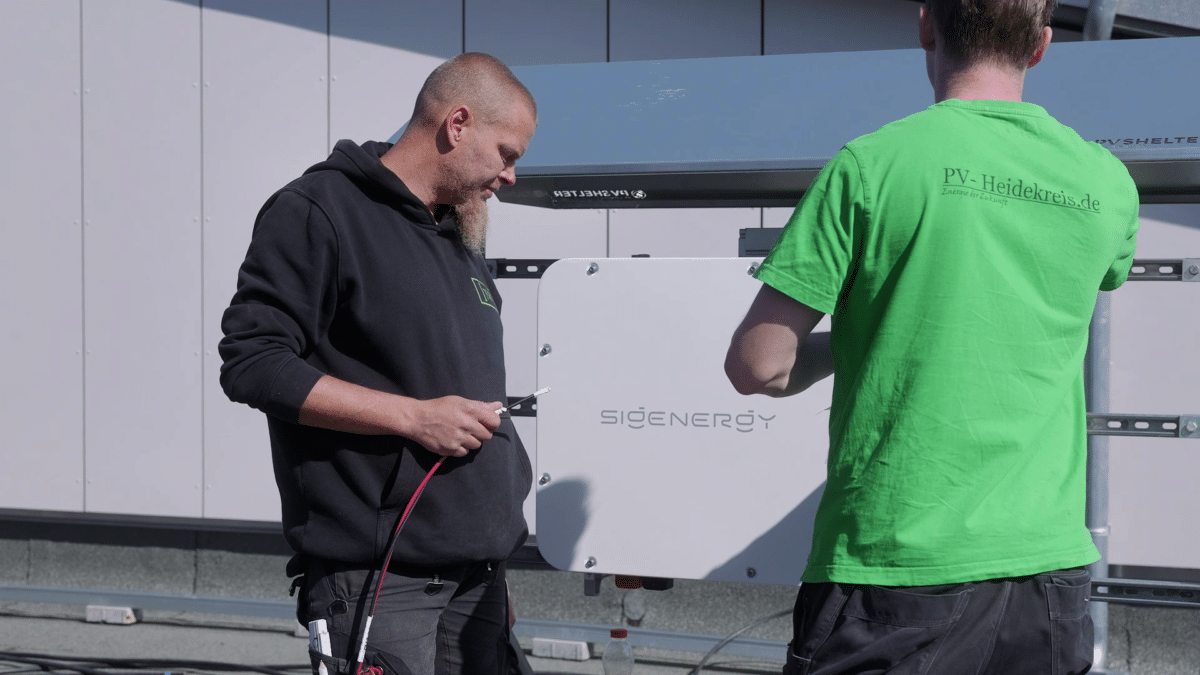 Zwei Monteure der INOL GmbH installieren einen Wechselrichter, der den Gleichstrom der Solarmodule in Wechselstrom umwandelt. Das weiße Gerät mit der Aufschrift Sigenergy hängt an einem Montagegestell vor einer grauen Fassade. Links hält ein Mitarbeiter mit Bart und schwarzem Kapuzenpullover ein rot-schwarzes Kabel, rechts arbeitet ein Kollege im grünen T-Shirt am Gerät. Elektroinstallation und Anschluss übernehmen bei INOL eigene Elektriker.
