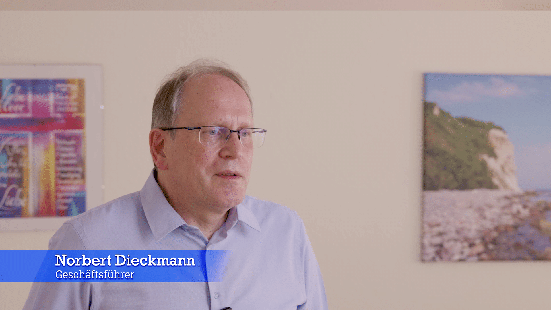 Norbert Dieckmann, Geschäftsführer der Stiftung Haus Zuflucht, im hellblauen Hemd vor einer hellen Wand mit zwei Bildern, unten eine blaue Einblendung mit Name und Funktion
